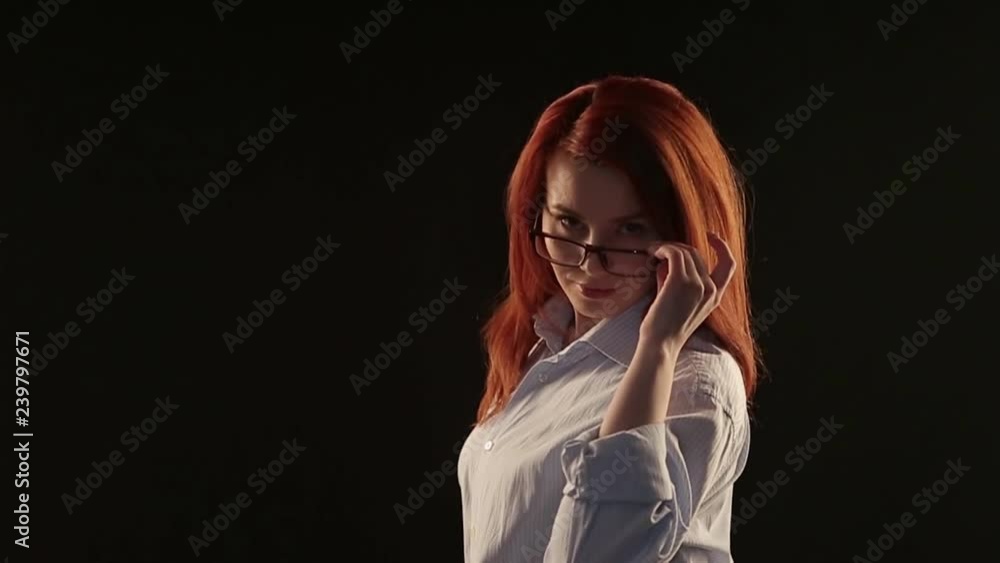 a beautiful girl with red hair in a white shirt posing on a black background in front of the camera. the model advertises glasses, lenses or services to restore vision a beautiful girl with red hair in a white shirt posing on a black background in front of the camera. the model advertises glasses, lenses or services to restore vision