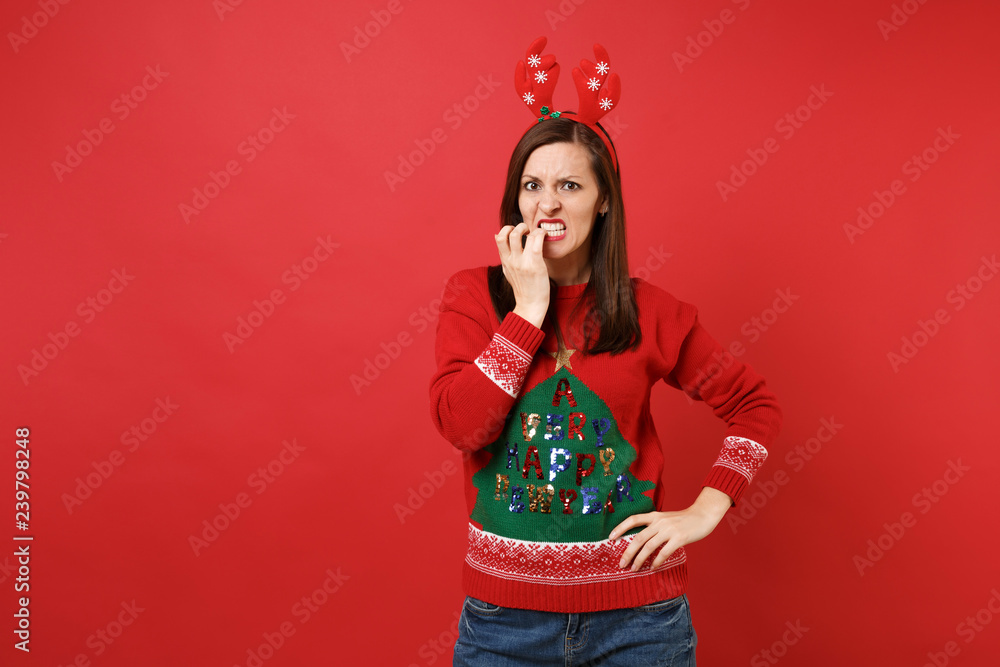 Concerned young Santa girl in knitted sweater fun decorative deer horns on head biting nails isolated on red wall background. Happy New Year 2019 celebration holiday party concept. Mock up copy space. Concerned young Santa girl in knitted sweater fun decorative deer horns on head biting nails isolated on red wall background. Happy New Year 2019 celebration holiday party concept. Mock up copy space.