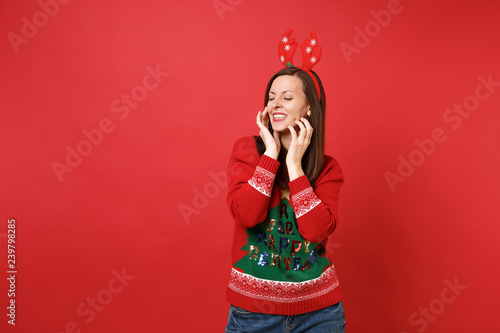 Tender young Santa girl in fun decorative deer horns keeping eyes closed, putting hands on face isolated on red background. Happy New Year 2019 celebration holiday party concept. Mock up copy space. © ViDi Studio