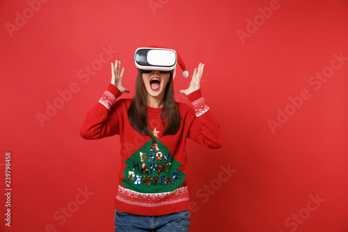 Screaming young Santa girl in knitted sweater, Christmas hat looking in headset, spreading hands isolated on red background. Happy New Year 2019 celebration holiday party concept. Mock up copy space.