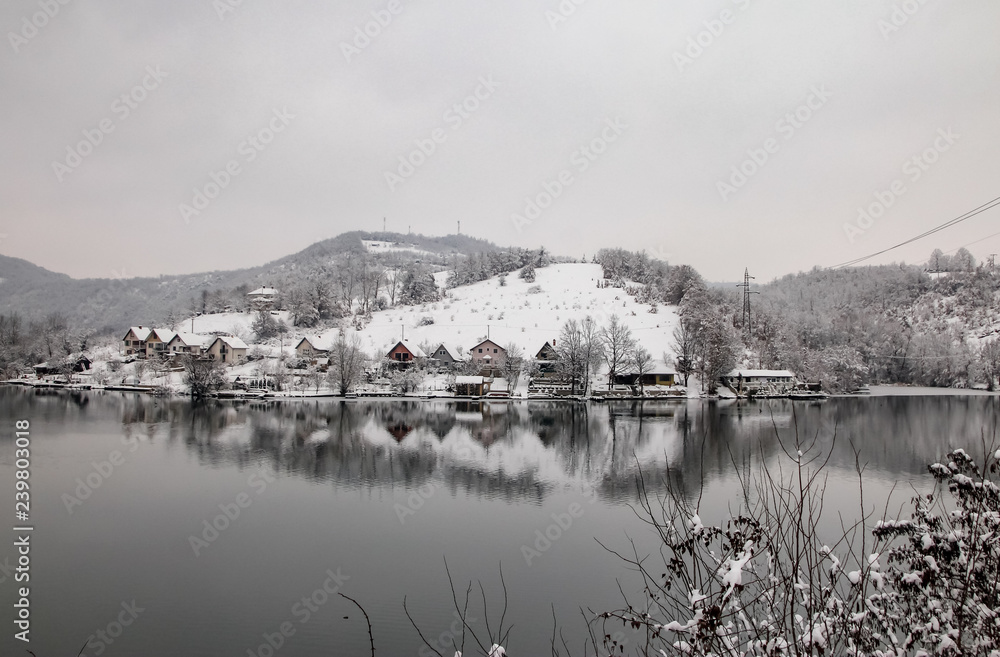 Houses near the frozen river covered with snow and surrounded by forest. Reflection in the water. River west Morava in Ovcar-Kablar gorge in Serbia.