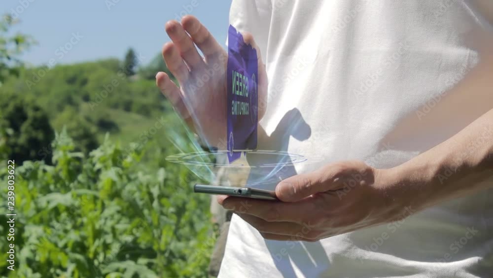Man shows concept hologram Green computing on his phone. Person in white t-shirt with future technology holographic screen and green nature background Man shows concept hologram Green computing on his phone. Person in white t-shirt with future technology holographic screen and green nature background