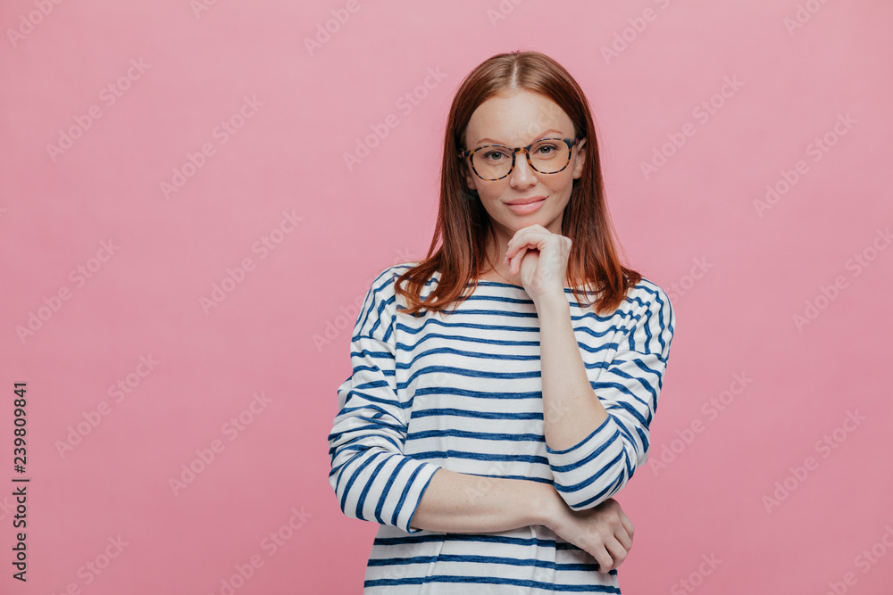 Indoor shot of attractive dark haired woman keeps hands under chin, wears striped sailor sweater, optical eyewear, models over pink background, looks directly at camera, has confident expression Indoor shot of attractive dark haired woman keeps hands under chin, wears striped sailor sweater, optical eyewear, models over pink background, looks directly at camera, has confident expression