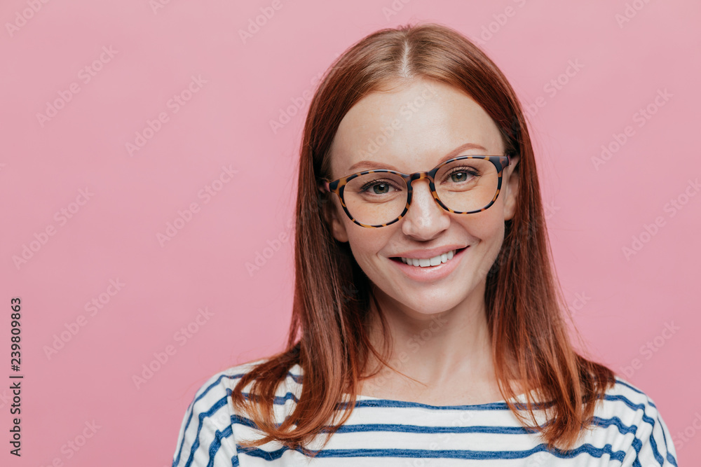 Close up shot of good looking brown haired woman with no make up, wears optical glasses, has gentle smile on face, dressed in striped clothes, models over pink background. Positive emotions concept Close up shot of good looking brown haired woman with no make up, wears optical glasses, has gentle smile on face, dressed in striped clothes, models over pink background. Positive emotions concept