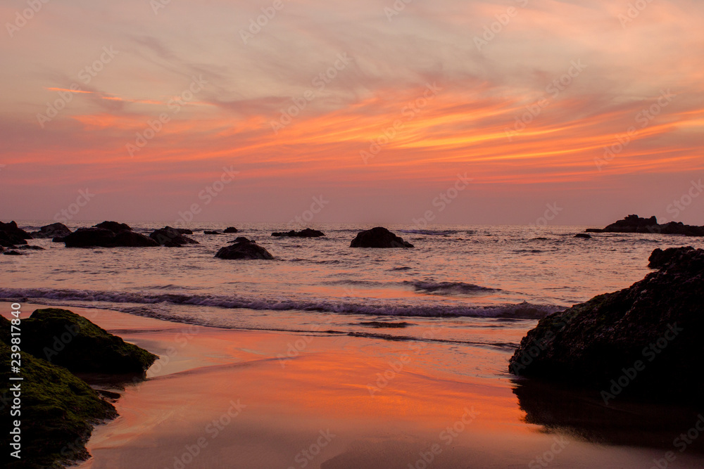 Brightly orange blue gray purple cloud lines in the sunset sky over the sea with rocks and a sandy beach