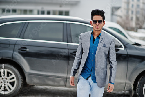 Casual young indian man in sunglasses posed against suv car.