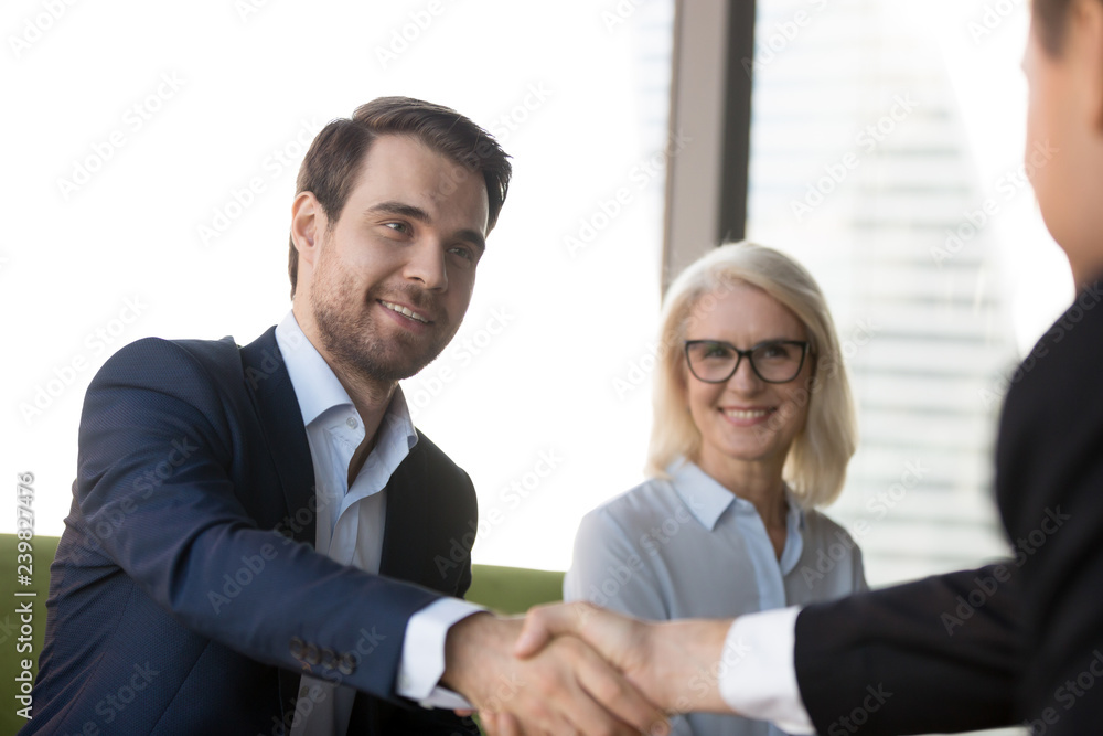 Business people greeting each other at meeting, focus on handsome young confident businessman welcoming client. Aged female in eyeglasses, team leader and company partner handshaking in modern office Business people greeting each other at meeting, focus on handsome young confident businessman welcoming client. Aged female in eyeglasses, team leader and company partner handshaking in modern office