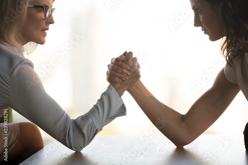 Side view close up young brunette and aged blond businesswomen arm wrestling holds elbows on table, exerting pressure on each other, looking eyes to eyes face to face struggling for leadership at work photo