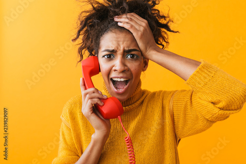 Portrait of young african american woman with afro hairstyle shouting and holding red handset, isolated over yellow background