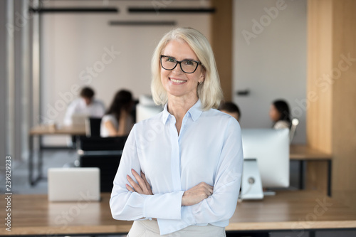 Confident executive manager middle aged female smiling looking at camera standing with crossed hands on chest in coworking space feels happy and satisfied. Team leader and successful employee concept photo