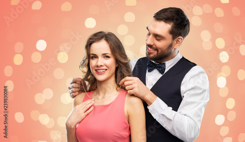 couple, jewelry and people concept - happy man puts necklace with diamond pendant on his girlfriend over living coral background and festive lights