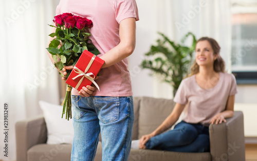 couple, relationships and people concept - happy woman looking at man hiding flowers and gift behind his back at home