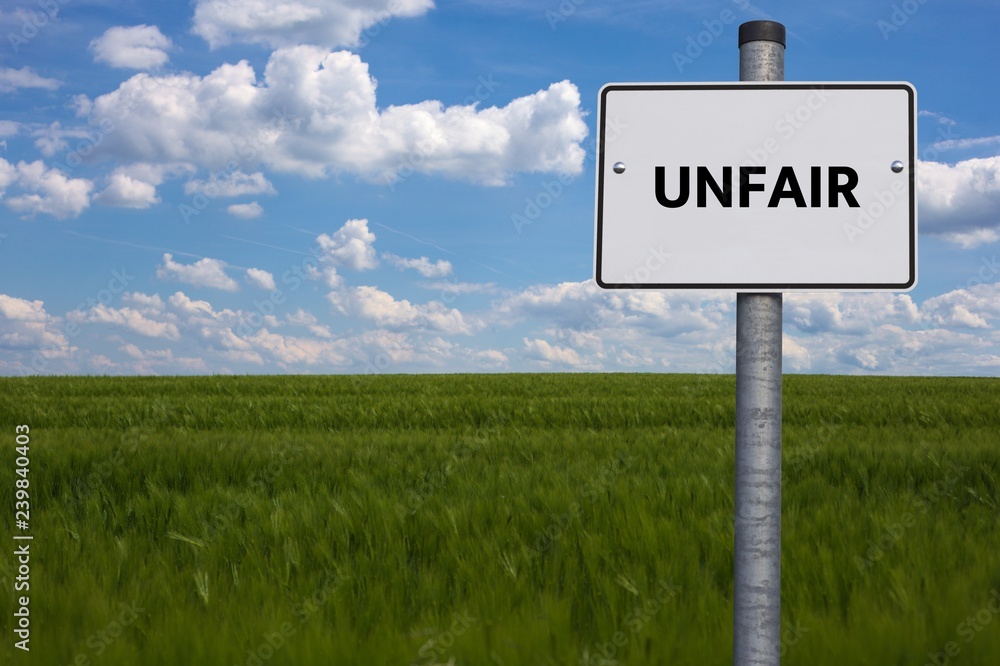 white road sign. the word UNFAIR is displayed. The sign stands on a field with blue background white road sign. the word UNFAIR is displayed. The sign stands on a field with blue background