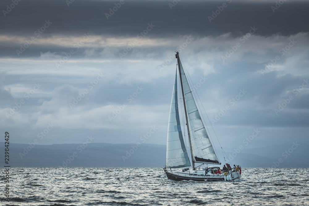 Lone touristic yacht sailing in the sea. Dark cloudy sky before the rain. Northern Ireland. Amazing marine scenery the small ship among the calm water. Ocean scape in grey blue tints.
