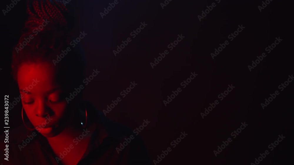 Panning low-key portrait shot with use of red color gel: confident black woman with hair bun and hoops earrings standing isolated on dark background and looking at camera with determination Panning low-key portrait shot with use of red color gel: confident black woman with hair bun and hoops earrings standing isolated on dark background and looking at camera with determination