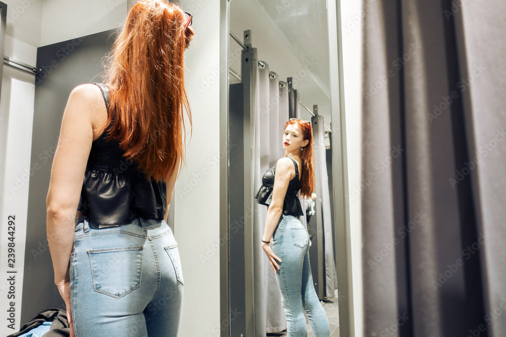young slim girl trying on new jeans in the fitting room in the store. woman  with beautiful figure looks at herself in the reflection of the mirror.  purchase foto de Stock