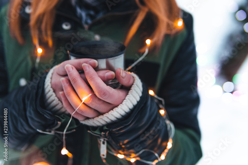 Close up view. Girl hands with a cup of hot coffee, tea, in the snowy forest. Concept adventure active vacations outdoor. Winter camping, copy space, colorful christmas lights