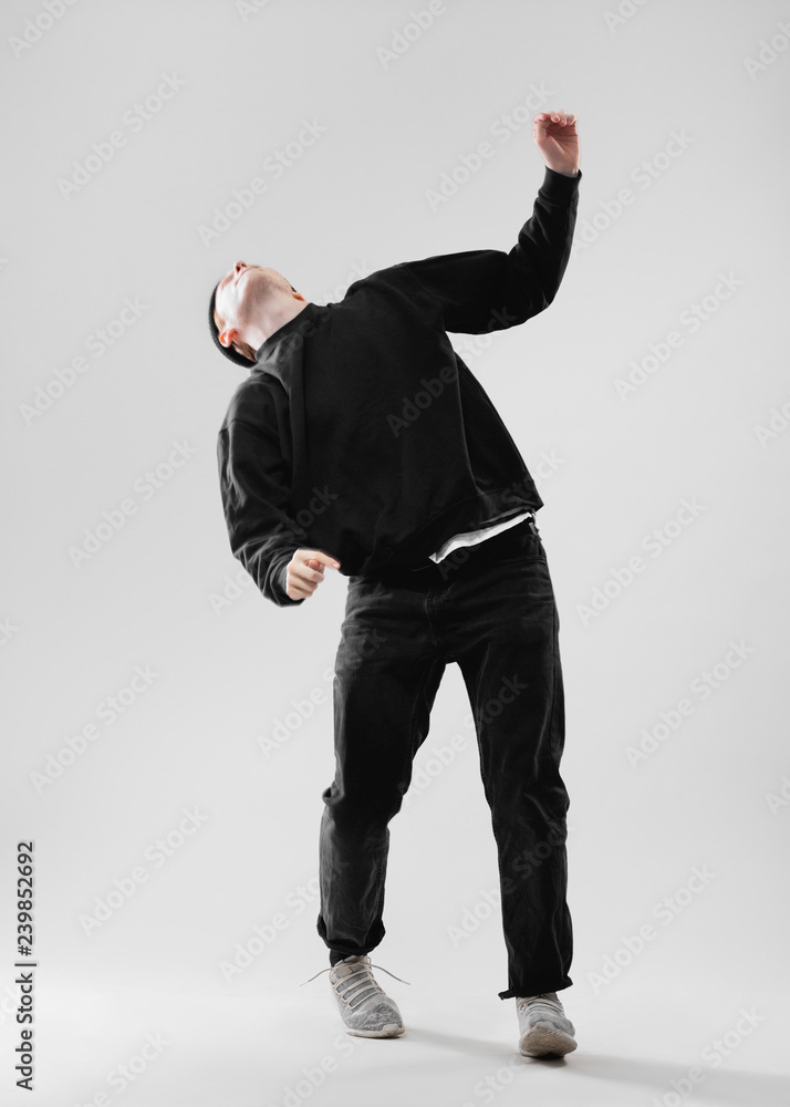 Dancer dressed in black jeans, sweatshirt, hat and gray sneakers is dancing  in the studio on the white background