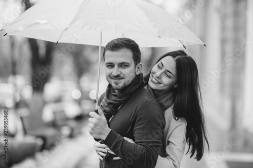 Happy romantic couple, guy and his girlfriend dressed in casual clothes are hugging under the umbrella and look at each other on the street in the rain. Black and white photo