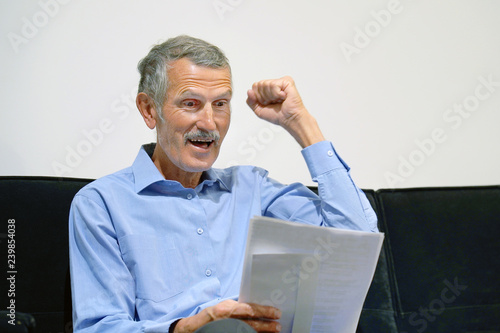 Elderly man enjoying good news in writing. The male reads a letter with good news sitting on the couch. An euphoric male is happy after reading good news in a written letter, approving a loan. photo