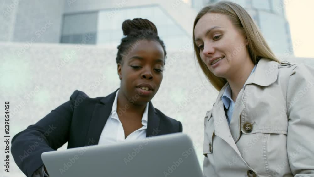 Panning of African and Caucasian female colleagues sitting together outdoors, looking at laptop screen and discussing details of business project Panning of African and Caucasian female colleagues sitting together outdoors, looking at laptop screen and discussing details of business project