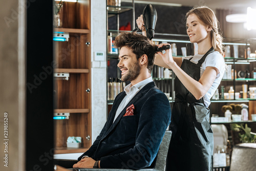 beautiful young hairdresser drying hair to handsome smiling man in beauty salon