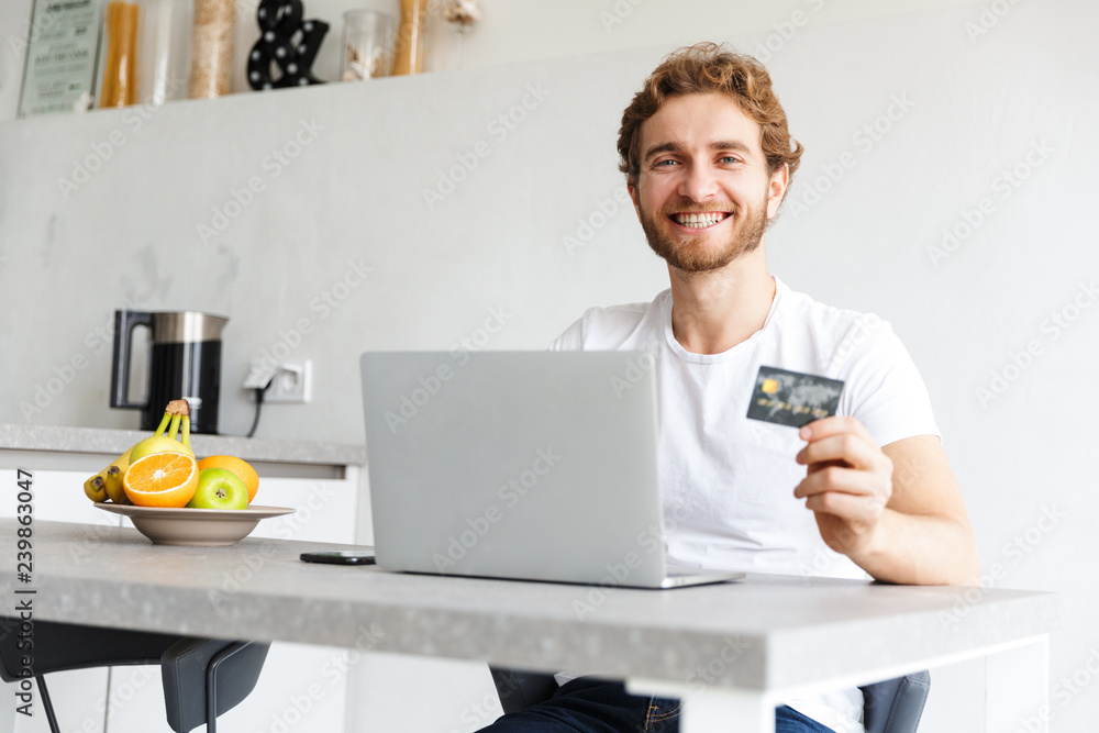 Happy young bearded man at the table at home talking using laptop computer holding credit card. Happy young bearded man at the table at home talking using laptop computer holding credit card.