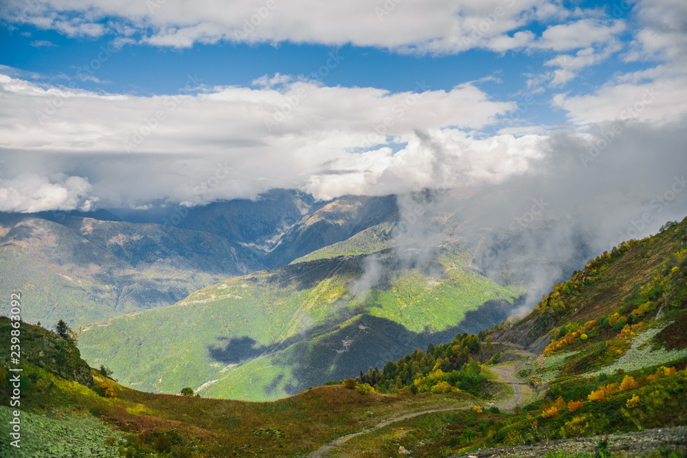 Mountain landscape. Warm autumn Sunny day. The slopes of the mountains covered with autumn woods. Clouds above mountain peaks. The snow on the tops Mountain landscape. Warm autumn Sunny day. The slopes of the mountains covered with autumn woods. Clouds above mountain peaks. The snow on the tops