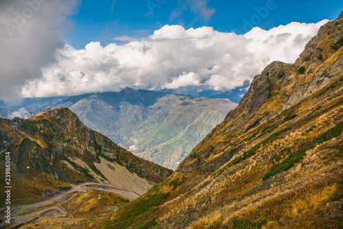 Mountain landscape. Warm autumn Sunny day. The slopes of the mountains covered with autumn woods. Clouds above mountain peaks. The snow on the tops