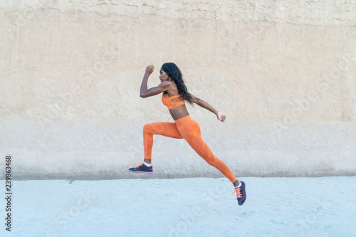 African American Black athletic model wearing bright orange sports outfit does a dramatic running leap against a concrete wall 