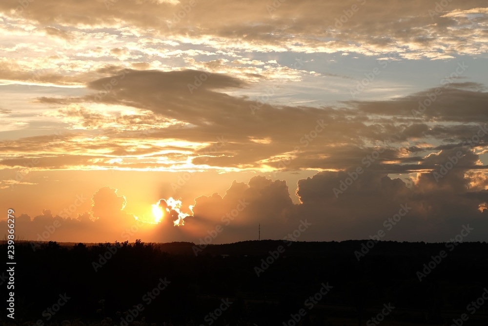 A Wide View Of An Orange / Red Coloured Sunset. The Sun Is Behind The Clouds And Causes A Bright Highlight Around Those Clouds. Landscape Can Be Seen In The Fore. Sunset Is Left Prioritized.