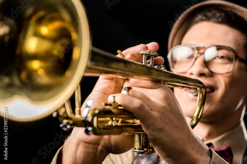 selective focus of male jazzman in hat and eyeglasses playing on trumpet isolated on black