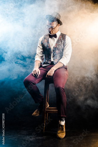 Fototapeta Naklejka Na Ścianę i Meble -  fashionable mixed race young man posing on chair on stage with smoke and dramatic lighting