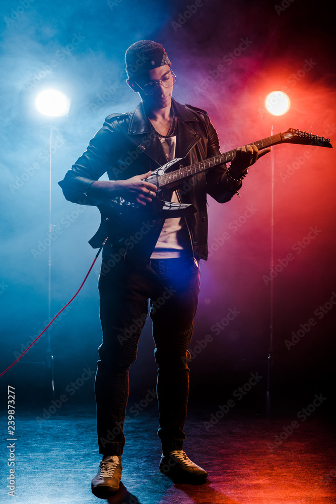 concentrated man in leather jacket performing on electric guitar on stage with smoke and dramatic lighting