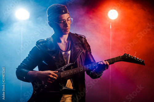 focused male musician in leather jacket performing on electric guitar on stage with smoke and dramatic lighting