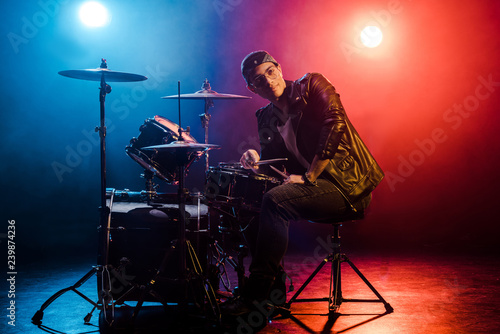 young mixed race male musician sitting behind drum set on stage with spotlights and smoke