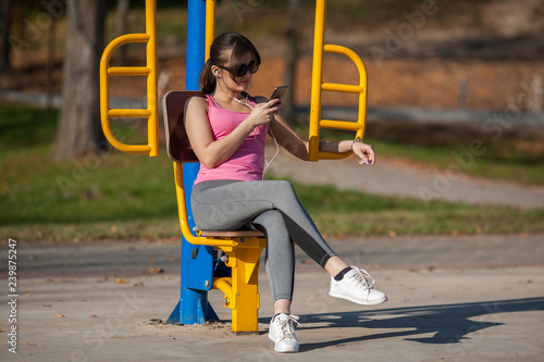 Young athletic girl in sunglasses is sitting on a sports simulator and listening to music on a street playground in the autumn.