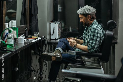 Calm man crossing legs while sitting with smartphone in barbershop