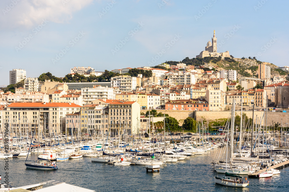 View over the Old Port of Marseille, France, with motorboats and sailboats moored in the marina and Notre-Dame de la Garde basilica on top of the hill at sunset.