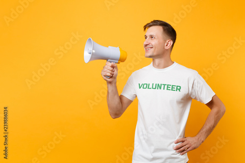 Portrait of man in white t-shirt written inscription green title volunteer scream in public address megaphone isolated on yellow background. Voluntary free assistance help, charity grace work concept.