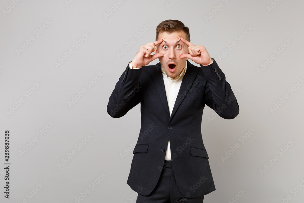 Shocked young business man in classic black suit keeping mouth wide open, stretching eyelids with fingers isolated on grey background. Achievement career wealth business concept. Mock up copy space. Shocked young business man in classic black suit keeping mouth wide open, stretching eyelids with fingers isolated on grey background. Achievement career wealth business concept. Mock up copy space.