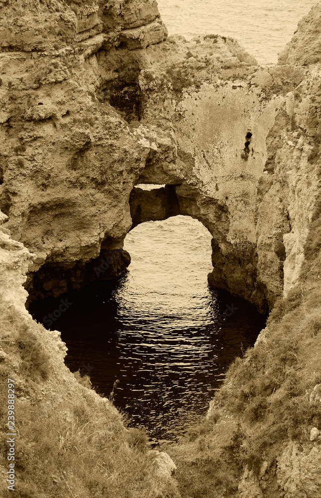 Ocean water seen through hole in rocks. Stone arches, caves, rock formations at Dona Ana Beach (Lagos, Algarve coast, Portugal) in the evening light. Sepia photo.