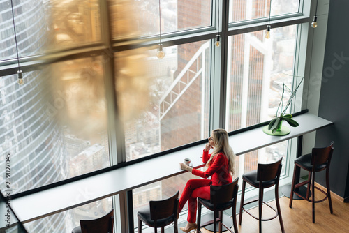Woman in red coral business suit drinks coffee at a high table near the window. Young blonde businesswoman in the office in high floor in front of the window. © malkovkosta