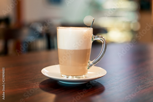 A cup of hot latte coffee in transparent glass on a wooden dark brown table and teaspoon in a cup with light bokeh background  front view.
