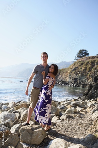 Young man and woman 20s smiling holding each other on rocky beach - wide shot
