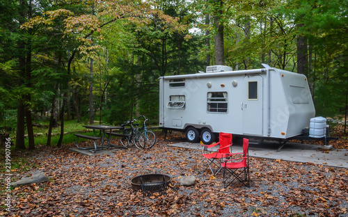 Camping in camper trailer in wooded park camp site with chairs set up around fire pit, and bicycles leaning against picnic table.