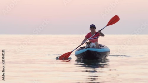 Varna, Bulgaria, mid December 2018. Young woman wearing a cap paddles on an inflatable kayak over the calm surface of the sea against the sky in the light of a pink dawn. Dog Jack Russell with kayak