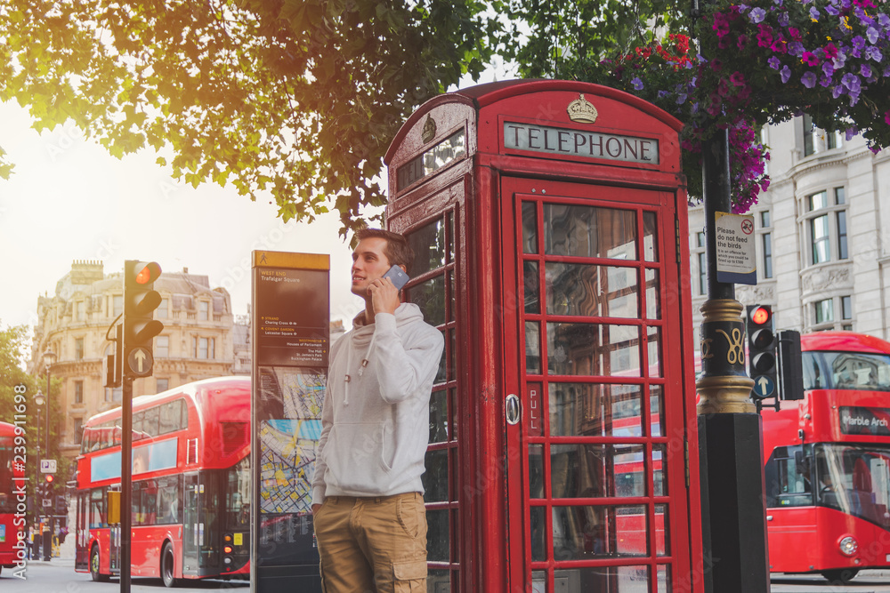 young boy using the smartphone in front of a phone box and a red bus in London - obrazy, fototapety, plakaty 