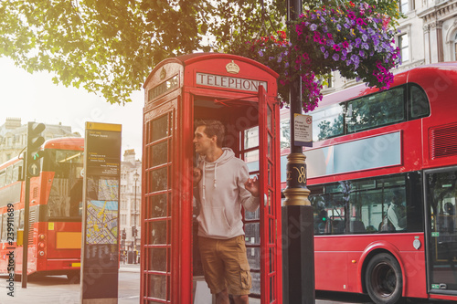 young male in London looking out from a phone booth with red busses in the back