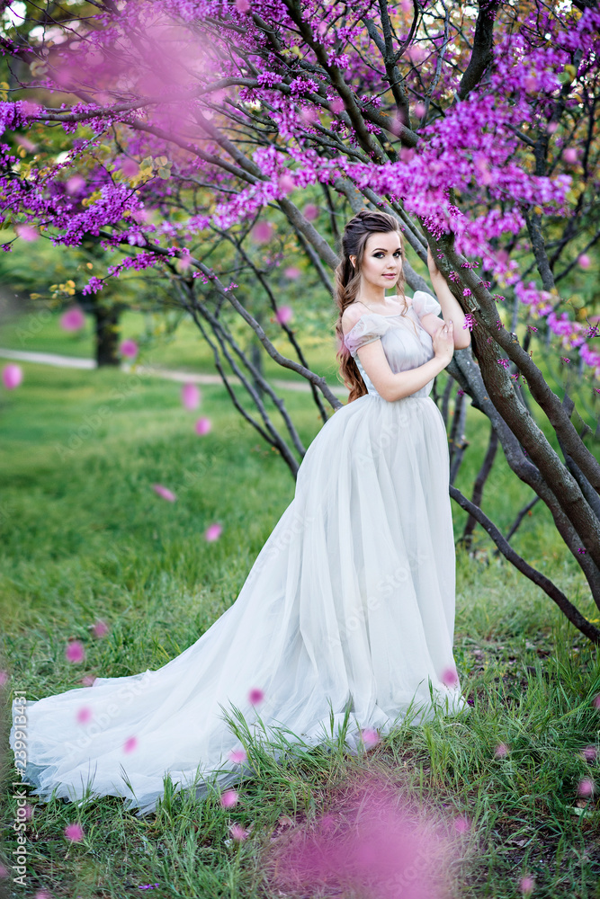 Luxurious and stylish bride poses in the garden among purple flowers. Happy bride with beautiful and long hair. An important day in life, strong emotions.